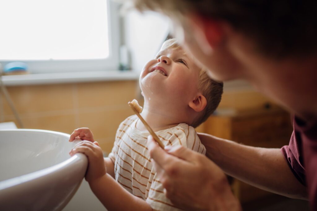 Man in red shirt helping little boy in striped t-shirt brush his teeth at bathroom sink
